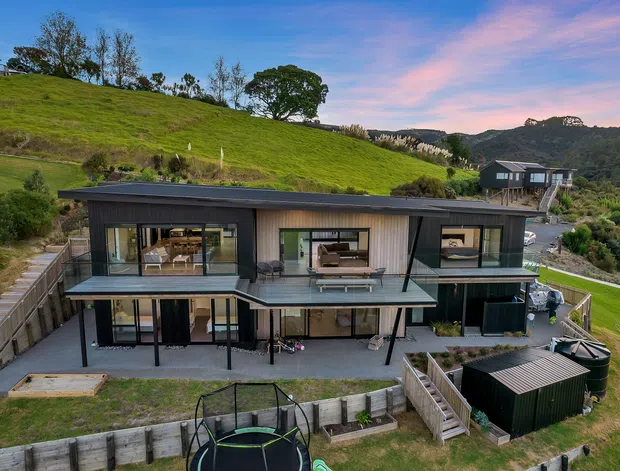 A contemporary multi-level house set into a sloping green site, viewed from above. The home has dark horizontal cladding, extensive floor-to-ceiling glazing, projecting upper-floor volumes, covered outdoor areas, timber retaining walls, and landscaped terraces. A smaller dark outbuilding and a trampoline are visible in the foreground, with rolling wooded hills beyond.