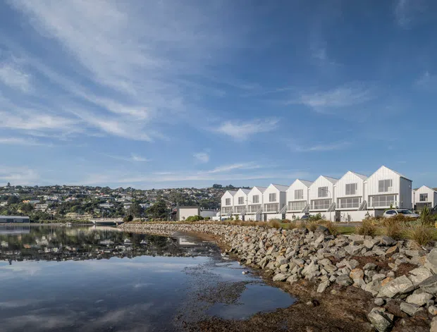 Exterior architectural view of a contemporary waterfront residential development, with a row of attached white gabled homes overlooking a calm inlet. The rocky shoreline, reflective water, distant suburban hillside, and expansive blue sky frame the buildings.
