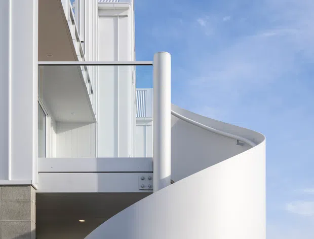 Close-up exterior architectural detail of a modern multi-storey building, featuring white rendered or painted curved balcony walls, slim metal balustrades, white vertical cladding or paneling, and a prominent cylindrical structural or drainage element. The composition emphasizes clean geometric forms against a blue sky.