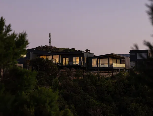 Twilight exterior view of a contemporary multi-level residential building set among dense vegetation on a sloped site. The structure features dark horizontal volumes, extensive illuminated glazing, projecting decks or terraces, and a low-profile flat or gently pitched roofline. A rooftop communications mast is visible, while foreground trees partially screen the building.