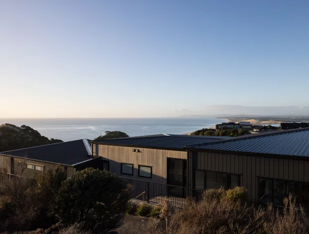 A contemporary single-storey coastal building with dark metal roofing and vertical timber-clad walls, viewed from a landscaped hillside. The low-profile structure overlooks the ocean, with scrubby native vegetation in the foreground and expansive sea and sky beyond.