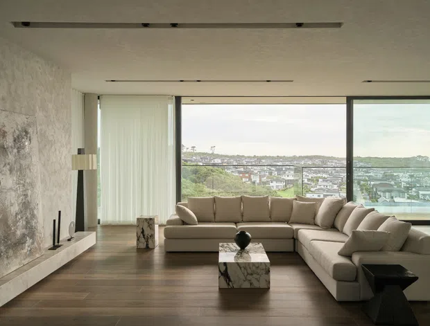 A refined contemporary living room with a large L-shaped beige sectional sofa arranged around a marble cube coffee table. Floor-to-ceiling windows provide expansive urban views, while a textured stone feature wall with a minimalist linear fireplace anchors the room. Dark timber flooring, sheer curtains, recessed linear lighting, and restrained neutral furnishings create a calm, luxurious interior.