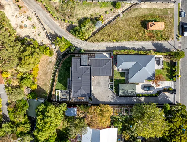 Aerial top-down view of a residential property on a landscaped, sloping suburban site. Several buildings with dark and light pitched roofs, paved driveways and paths, retaining walls, gardens, mature trees, and adjacent streets are visible.