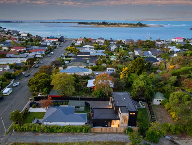 Aerial view of a suburban coastal neighbourhood beside a broad sheltered bay, with a modern dark-clad residence in the foreground, pitched and flat roof forms, tree-lined streets, gardens, and low-density housing extending toward the water. Several small islands or headlands and boats are visible across the bay.