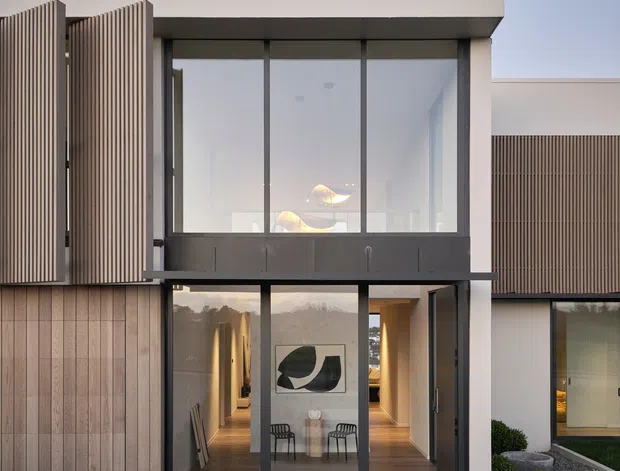 Exterior view of a contemporary multi-level residence with a rectilinear form, broad flat roofline, dark-framed floor-to-ceiling glazing, and vertical timber screening. A stepped concrete path crosses a dark gravel landscape toward a glazed entry, with interior furnishings visible through the windows.
