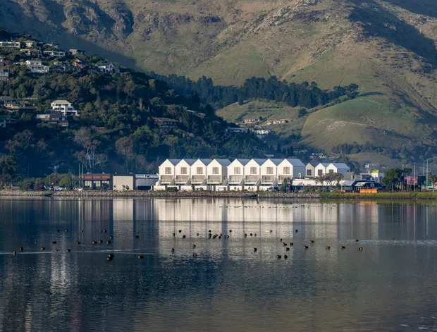 A waterfront architectural exterior viewed across calm water, showing a long row of attached white buildings with repeated steep gabled roof forms and dark window openings. Hillside homes and rugged mountains rise behind the development, while the water creates a clear reflection in the foreground.
