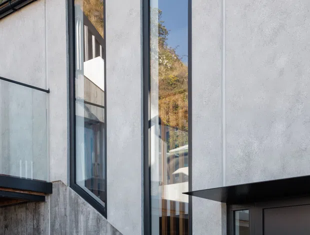Close exterior view of a contemporary multi-level facade on a sloping site. The building has white textured rendered walls, tall dark-framed windows, black gutters and fascia, a dark entrance canopy, and a lower raw concrete retaining wall. Reflections reveal interior glazing and structural elements, while surrounding vegetation and hillside are visible in the background.