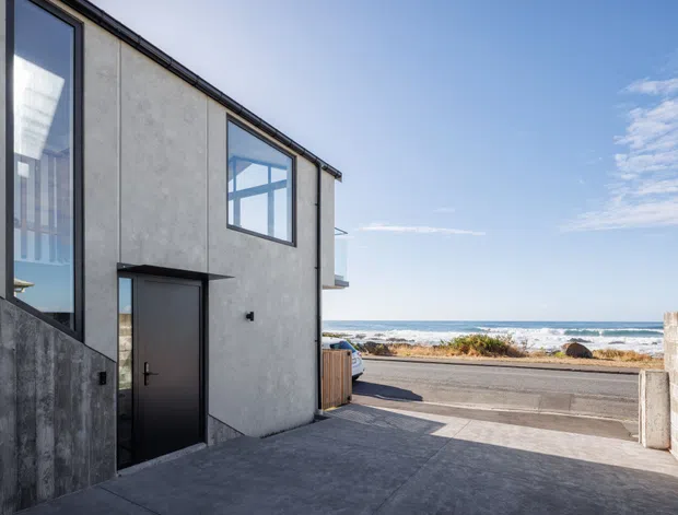 Exterior view of a contemporary coastal residence, showing a textured pale rendered facade with dark metal-framed glazing and a dark entry door. The driveway slopes toward a road and a visible ocean shoreline beyond, with a low masonry boundary wall at right.