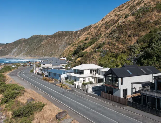 Aerial or elevated view of a coastal residential settlement beside a winding road, with modern detached homes clustered between the ocean and steep rugged hills. The scene includes dark pitched roofs, light-coloured facades, balconies, and a dramatic coastal landscape under clear blue sky.
