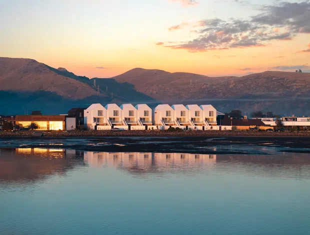 Exterior architectural view of a contemporary waterfront development, featuring a row of identical white gabled multi-storey units beside calm reflective water. Low commercial or service buildings extend to either side, with rugged mountainous terrain in the background and warm sunset light.