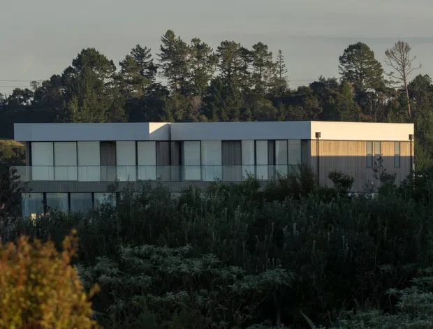 A modern, low-profile two-storey residence viewed from across dense vegetation. The building has a rectilinear flat-roof form, light-coloured rendered or panelled walls, extensive floor-to-ceiling glazing with dark slender frames, and a projecting upper-level volume. Mature trees and landscaped planting surround the property in soft, subdued evening light.