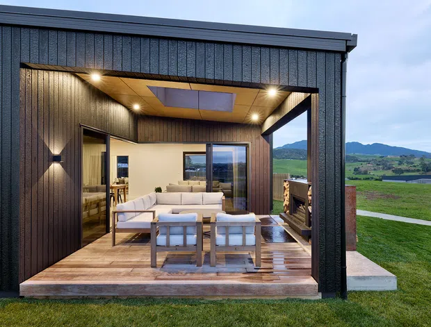 Exterior view of a compact contemporary pavilion or living extension set in a rural landscape. Dark vertically textured cladding frames an open covered terrace with timber decking, built-in warm lighting, and an outdoor seating arrangement. Through the opening, a bright living room with a sofa, armchairs, timber flooring, and glazing is visible. Rolling green farmland and hills extend beyond the structure.