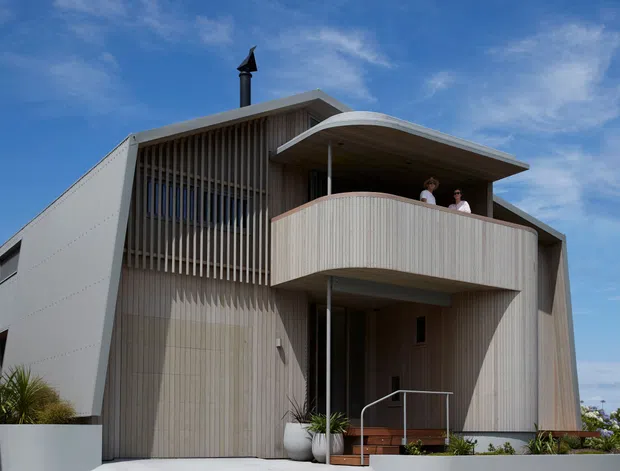 Exterior view of a contemporary multi-level residence with a sculptural curved balcony canopy and rounded projecting balcony. The facade combines pale vertical timber-like screening and cladding with white angular wall planes, recessed glazing, a dark flue, and a small entry stair. Coastal planting and a bright blue sky surround the building.