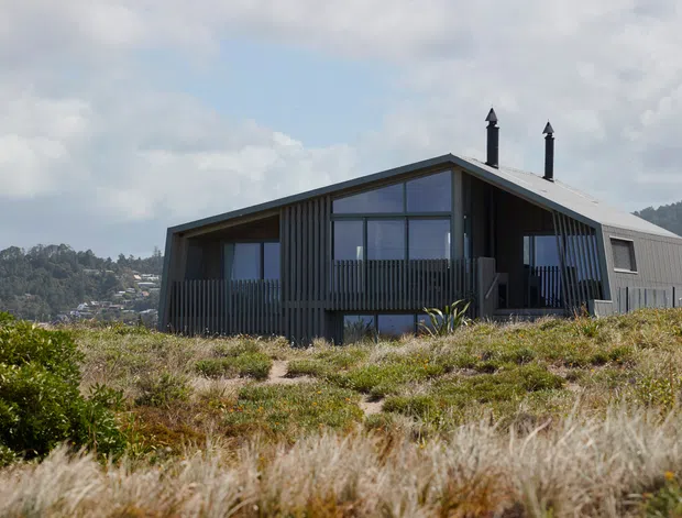 A contemporary single-storey dwelling set in a grassy coastal or elevated landscape. The house has a steep asymmetrical gable roof, dark corrugated metal cladding, extensive floor-to-ceiling glazing, and a covered front deck with vertical timber or metal balustrading. Two chimney flues project through the roof. Hillside housing and bush-covered terrain are visible in the background.