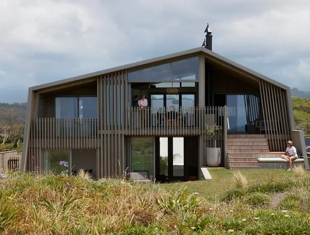 Exterior view of a contemporary two-storey house set in a landscaped rural or semi-rural site. The home has a steep asymmetrical gable roof, extensive glazing, dark vertical timber-like cladding and screening, a projecting upper balcony with vertical balustrades, and an external stair leading to the upper level. People are present on the balcony and stairs, with hills and vegetation in the background.