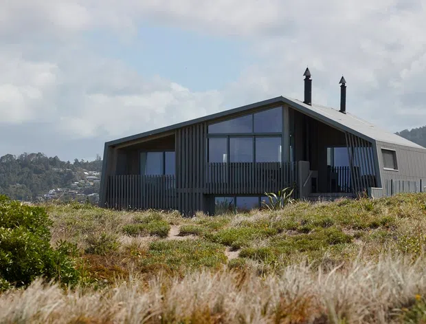 A contemporary dark-clad house set within coastal scrub or dune vegetation. The single-storey dwelling has a steep pitched gable roof, broad glazed openings, a covered recessed veranda, vertical timber screening and two slim chimney flues. A wooded hillside and scattered buildings are visible in the background.