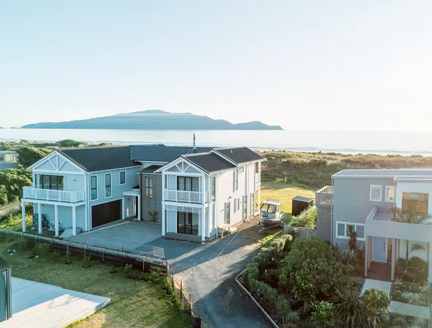 Aerial exterior view of a coastal residential development with several light-coloured multi-storey houses, dark pitched roofs, balconies, decks, and large glazed openings. The homes sit beside sandy coastal vegetation with ocean water and a distant island or headland visible beyond.