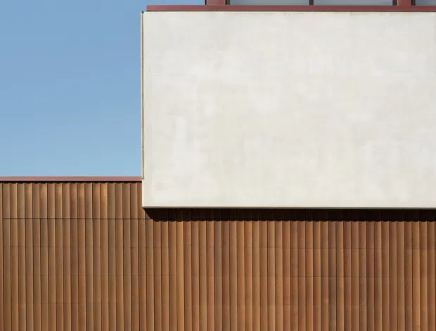 Minimalist architectural exterior with a large smooth off-white rendered upper volume projecting over a warm brown vertically ribbed facade. A narrow dark-framed horizontal window and reddish metal roof or parapet trim are visible above, while pale rectangular pavers form the foreground. Strong sunlight and tree shadows create a clean, graphic composition against a clear blue sky.