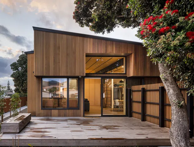 Exterior view of a contemporary two-storey home with a warm vertical timber-clad facade, large dark-framed glazed openings, and a recessed upper-level window. A broad timber deck forms the foreground, with a slatted privacy gate and mature flowering tree framing the right side. Warm interior lighting is visible through the glazing.