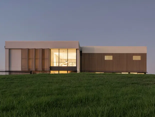 A low, single-storey contemporary building viewed from across a grassy site at dusk. The composition features two connected rectilinear volumes: a pale flat-roofed central section with warm illuminated full-height glazing and vertical timber-like screening, alongside a darker brown horizontally elongated volume with small high-level windows. The building sits on a gently elevated grassy landscape against an open evening sky.