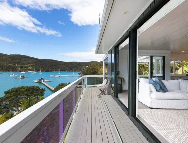 A bright coastal home viewed from a spacious timber-look balcony overlooking a turquoise bay with boats and wooded hills. Large black-framed sliding glass doors open into a relaxed white living room with blue cushions, light carpet, and a white ceiling. The balcony has a white balustrade, outdoor seating, and expansive waterfront views.