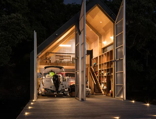 Night-time exterior photograph of a compact, elevated timber-clad structure set among dense trees. Large open bi-fold or hinged doors reveal a warmly lit, double-height interior with a steep gabled timber ceiling, mezzanine-style railing, extensive open shelving, and a boat or recreational vehicle stored inside. A broad timber deck with integrated step and floor lighting leads up to the building.