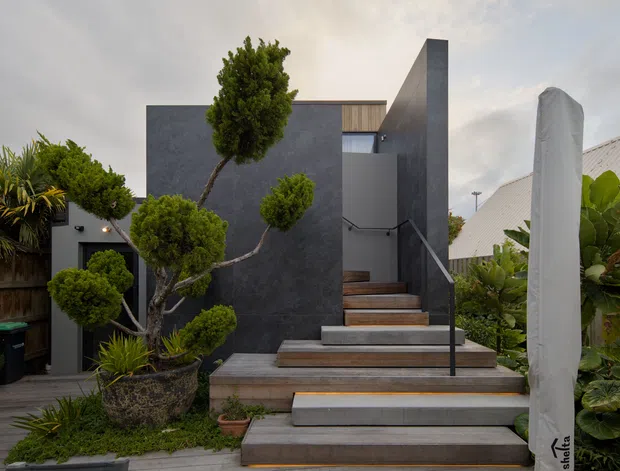 Exterior view of a modern multi-level residence, featuring dark textured wall cladding, a broad timber-and-concrete stairway with slender black handrails, and sculpted garden planting. The entrance is partially screened by a tall covered outdoor object, with neighbouring roof forms visible behind the landscaped boundary.