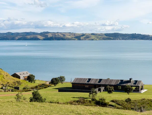 A wide exterior landscape view of a contemporary low-slung residence set on a gently sloping grassy coastal property, overlooking a broad calm bay and rolling hills. The building has dark timber-like cladding, a long linear form, and multiple pitched roof planes with skylight-like openings. A smaller outbuilding sits nearby, with expansive lawn and scattered native-looking vegetation in the foreground.