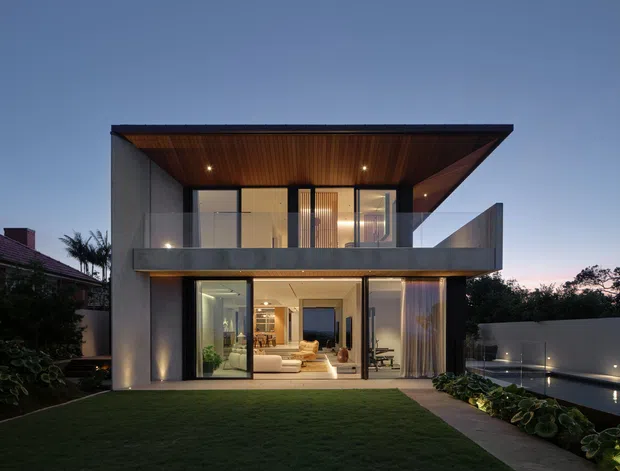 Twilight rear exterior view of a contemporary two-storey residence with a strong projecting flat roof, broad glazing, dark-framed windows and doors, a warm timber-lined soffit, and an upper-level balcony. The ground floor opens toward a landscaped lawn and a partially visible pool or water feature, while warm interior lighting reveals living spaces inside.