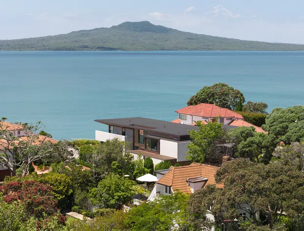 A sunny elevated view across a leafy coastal residential neighbourhood toward vivid turquoise water and a forested island or headland. Several homes are visible among dense trees, including a contemporary flat-roofed house with dark roof cladding and light walls, alongside traditional homes with terracotta tiled roofs.