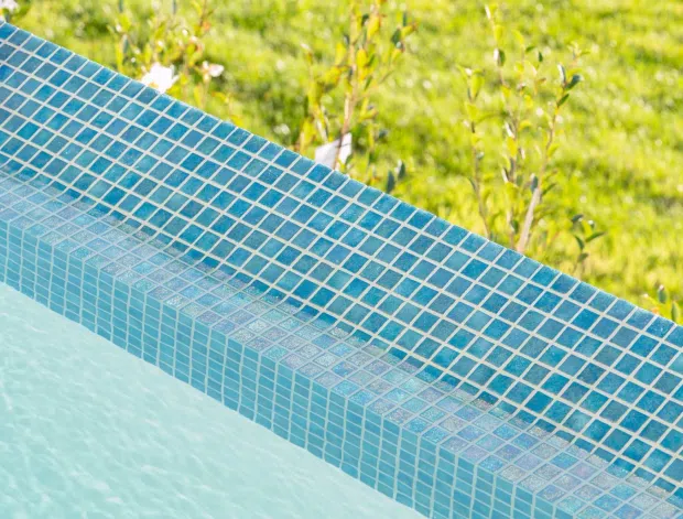 Close-up detail of a swimming pool edge lined with small square turquoise-blue mosaic tiles. The curved coping and waterline are visible, with a blurred green lawn and planting beyond the pool.