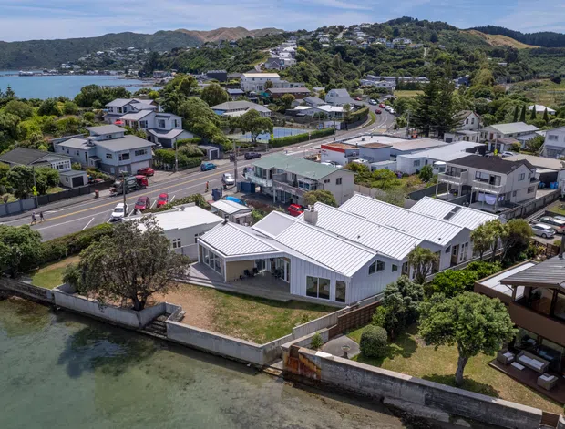 Aerial view of a coastal suburban neighbourhood beside a sheltered tidal waterfront. The foreground shows a modern single-storey residential property with light metal roofing, pale walls, glazing, landscaped gardens and a seawall. Roads, detached homes, hillside housing and vegetated coastal terrain extend into the background.
