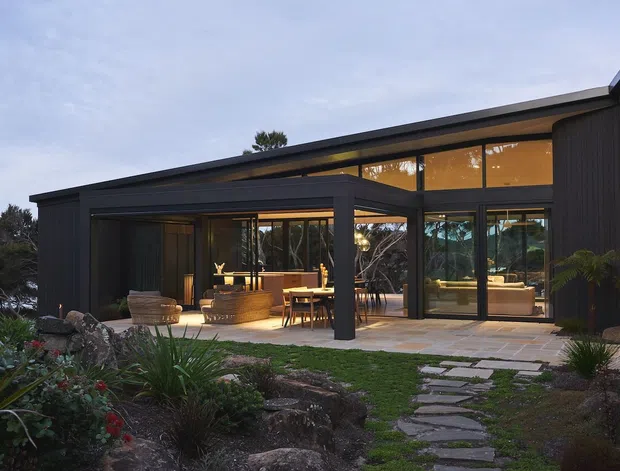 Exterior view of a contemporary dark-clad single-storey home with a low-pitched roof, extensive floor-to-ceiling glazing, and a sheltered outdoor entertaining area. Warm interior lighting reveals an open-plan living and dining space through the glass. A paved stepping-stone path leads through landscaped planting and rocks toward the house.