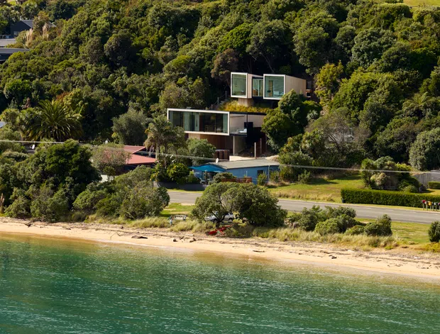 A long-distance exterior view of a modern multi-level home set into a densely vegetated hillside above a sandy waterfront. The house has rectilinear volumes, extensive floor-to-ceiling glazing, dark and neutral exterior surfaces, and a blue swimming pool or water feature near the road. The foreground shows calm green-blue water and a sandy shoreline.