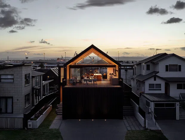 Twilight exterior view of a compact multi-storey residential building in a dense suburban setting. The central dark-clad home features a strongly expressed pitched gable roof, a warm timber-framed glazed upper-level living space with balcony railing, and a darker lower facade. Neighbouring houses, driveways, street lighting and a distant coastal-looking horizon are visible in the background.