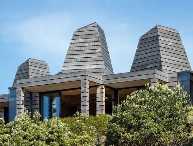 Exterior view of a contemporary, low-rise residence with three prominent tapered chimney-like tower forms and extensive horizontal grey timber cladding. Deep roof overhangs and recessed glazed openings create sheltered outdoor-facing spaces, while dense native-looking shrubs partially screen the lower facade. The composition is set against a clear blue sky.