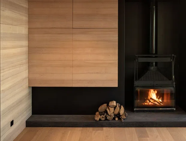A refined contemporary fireplace feature wall with warm timber wall panelling and cabinetry above a dark stone hearth. A black wood-burning fireplace with a visible flame and stacked firewood creates a strong focal point, while timber flooring and dark wall surfaces add warmth and contrast.