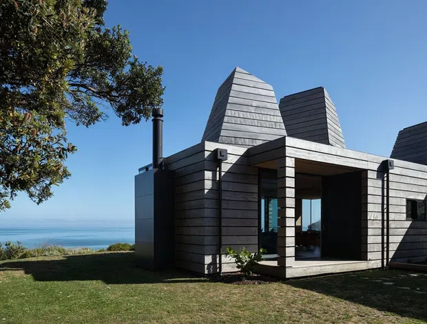 A modern coastal house viewed from the lawn, clad in horizontal grey weatherboards with several distinctive steep, faceted roof forms. Deep recessed openings and large glazed doors create a sheltered outdoor-facing entry or living area. The property sits beside a broad blue coastal or ocean outlook, with mature landscaping and strong clear daylight.