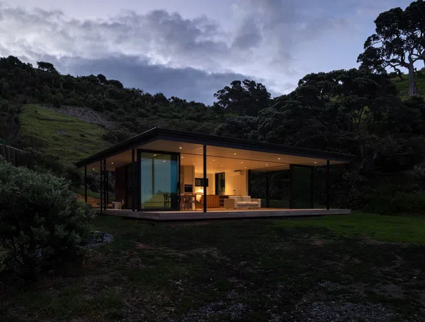 A low, minimalist single-storey pavilion set into a lush, steeply sloped landscape. The building has extensive floor-to-ceiling glazing, slender dark window frames, a broad shallow roof, and warm interior lighting visible from outside. A simple furnished living area is visible through the glass, while dense vegetation and rolling terrain surround the structure at dusk.