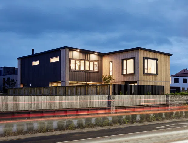 Twilight exterior view of a contemporary two-storey dwelling with a strong rectangular form, dark vertical cladding on one section, lighter timber-toned vertical cladding on the other, large framed windows, and a timber privacy fence. The wet roadway and vehicle light trails emphasize an urban suburban setting.