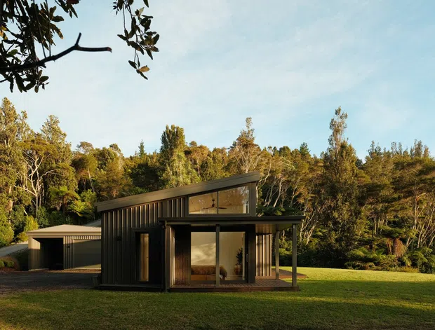 A small contemporary dark-clad pavilion or house set within a broad lawn and dense woodland. The building has a low asymmetrical pitched roof, extensive glazing, vertical ribbed wall cladding, and a separate matching outbuilding or carport to the left. Warm low-angle light highlights the timbered landscape setting.