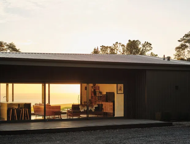 Single-storey contemporary home with a long, low-profile form, dark vertically ribbed exterior cladding, and a shallow pitched metal roof. Large glazed sliding doors reveal a warm living and dining interior, while a broad dark timber deck fronts the building. The home is set in a natural, semi-rural landscape and appears to overlook a golden coastal or open landscape.