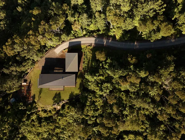 Aerial view of a dark-roofed, angular residential building set within dense forest, with a curved access road and small landscaped clearing visible around the house.