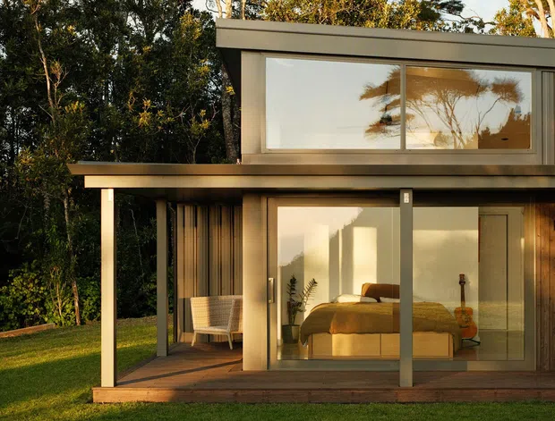 Exterior view of a compact two-storey modern home set among mature trees and lawn. The building has a flat roof, strong horizontal forms, extensive floor-to-ceiling glazing, dark slender framing, timber wall cladding, and a covered ground-floor terrace. A bedroom with a bed and bedside table is visible through the glazing.