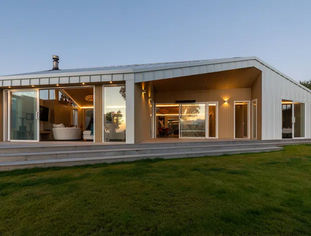 Single-storey contemporary residential house viewed from the lawn, with a long low-slung form, shallow pitched roof, extensive glazing, and a covered recessed outdoor living area. The exterior uses pale vertical metal cladding and timber-lined soffits, with warm interior and exterior lighting visible through open and glazed doors. A living room is visible at the left and a dining or living area behind the central glazing.