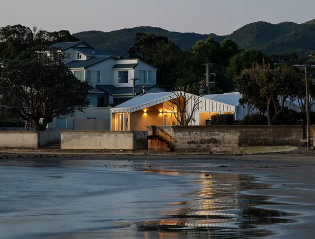 A dusk exterior view of a contemporary coastal residential property, seen across shallow reflective water. The home combines light-coloured rendered walls with blue-grey gabled roof forms and a low metal-clad structure with warm illuminated glazing. A concrete boundary wall fronts the property, with wooded hills rising behind it.