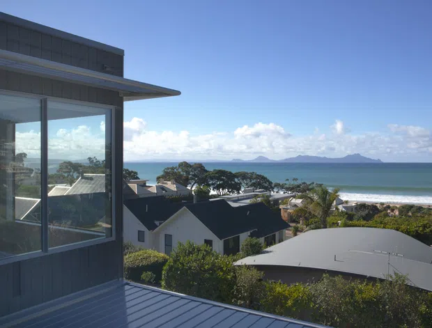 Exterior architectural view from an elevated residential building, featuring dark vertical cladding, extensive corner glazing, a projecting roof or canopy, and a metal-clad deck or balcony. The view overlooks a coastal suburban neighbourhood, sandy beach, ocean, and distant islands.
