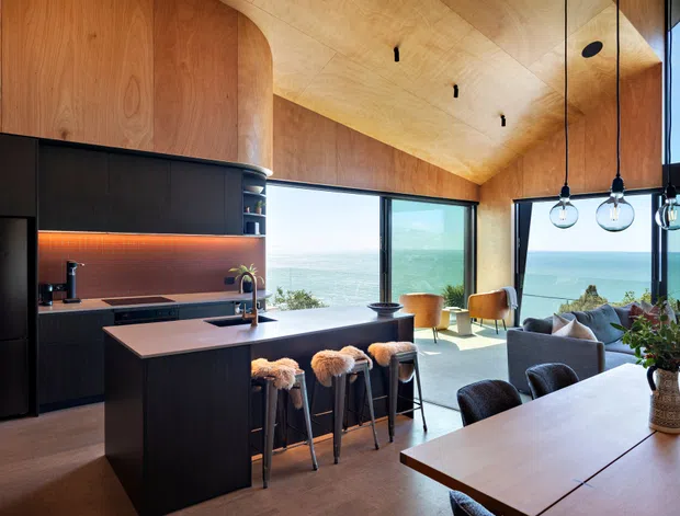 Open-plan contemporary kitchen, dining, and living area with a dramatic timber-lined vaulted ceiling and curved timber cabinetry. The kitchen features dark built-in cabinetry, a light stone island benchtop, integrated appliances, and warm under-cabinet lighting. A large dining table and pendant lights occupy the foreground, while full-height glazing frames expansive ocean and coastal views. The room is bright, warm, and refined, with a strong connection to the landscape.