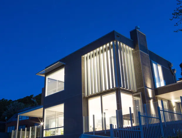 Dusk exterior view of a contemporary multi-level house with dark cladding, extensive illuminated glazing, prominent vertical sun-shading fins, a tall chimney, and a covered entry or terrace area behind a metal fence.
