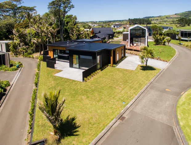 Aerial view of a contemporary residential development with a low-profile dark-clad house on a landscaped corner lot. The home features a flat roof, extensive glazing, dark exterior cladding, and a small paved driveway, surrounded by manicured lawns, palm-like plantings, and neighbouring modern homes on gently rolling terrain.